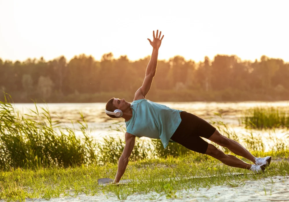 En mann trener utendørs ved en innsjø ved solnedgang og gjør en sideplanke. trening for depresjon kan øke mental helse. Besøk psykiater.no.