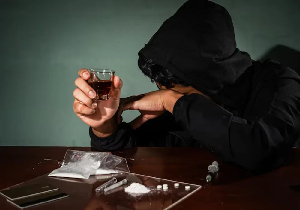 En person i hettegenser med alkohol og narkotika foran seg – et bilde som illustrerer sammenhengen mellom narkotikamisbruk og kriminalitet.