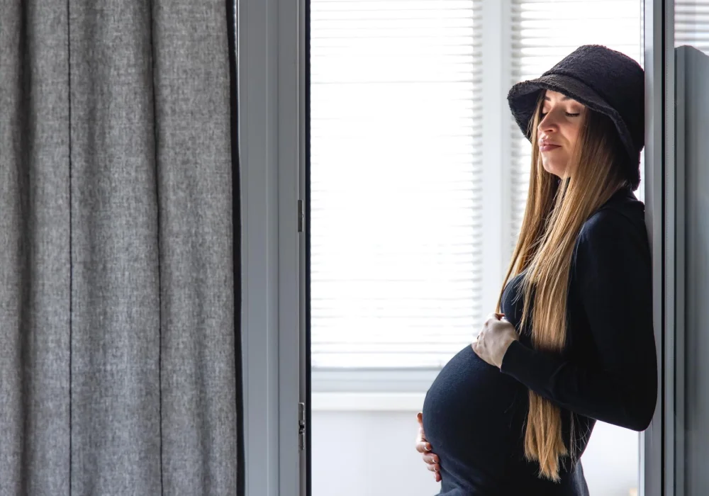 En gravid kvinne i svart klær og hatt står ved vinduet med øynene lukket og hånden på magen – illustrerer temaet depresjon-og-graviditet.